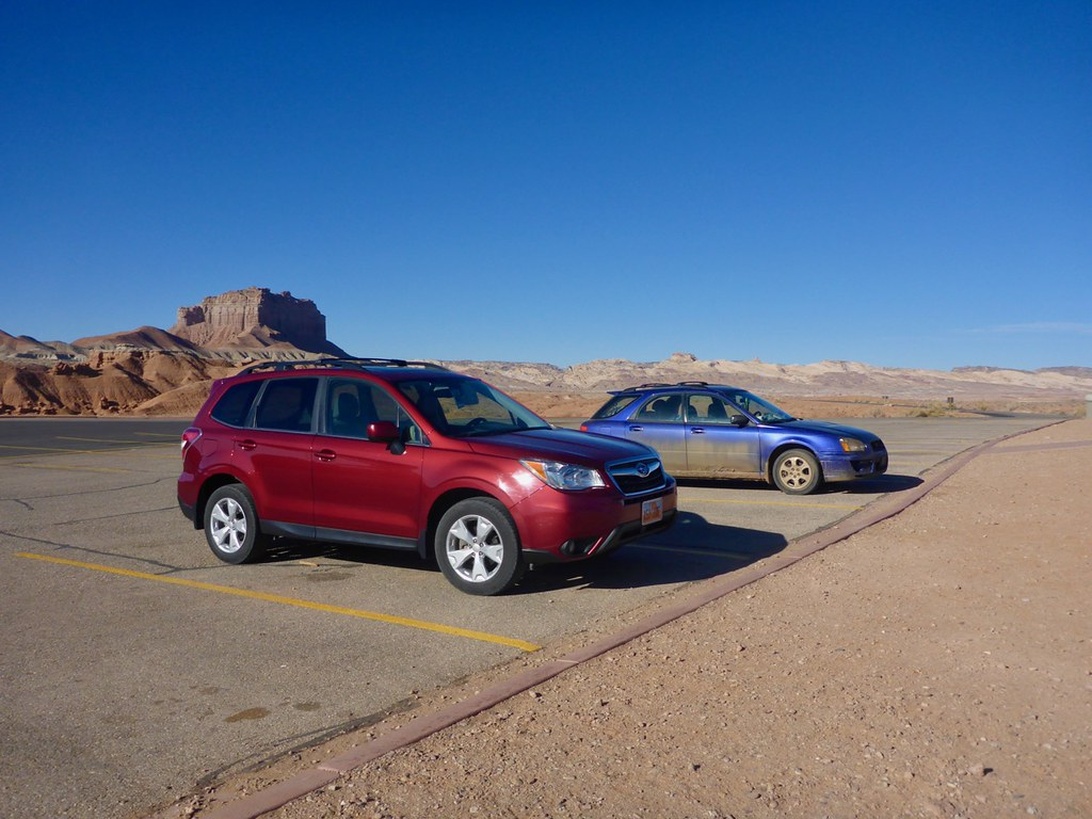Un SUV rouge et une berline bleue garés sur un parking dans un paysage désertique avec des formations de mesas et des montagnes en arrière-plan, évoquant le sud-ouest américain.